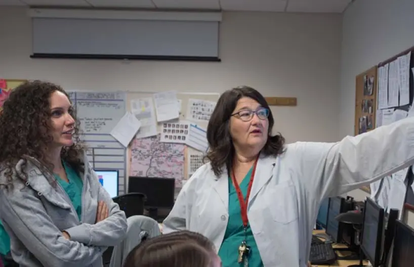 Two women, dressed in medical scrubs, engage in a discussion at a workspace filled with computers and bulletin boards.