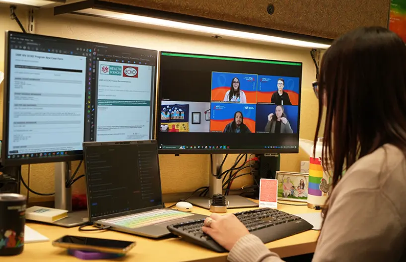 A person sitting at a desk with multiple monitors, engaged in a video conference, displaying various content and participants.