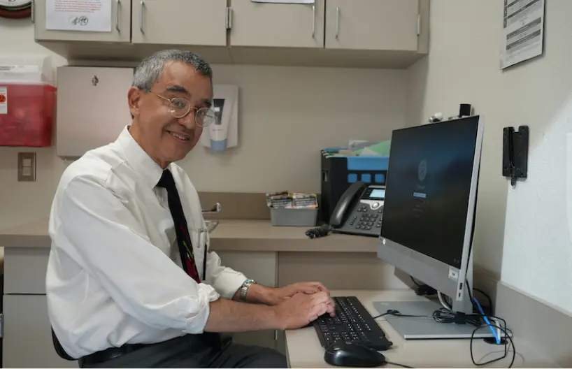 A smiling man in a white shirt and tie works at a desk with a computer and office equipment in a professional setting.